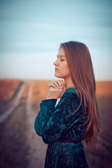 Young girl with long hair in a green dress. Standing on a country road near the field, with eyes...
