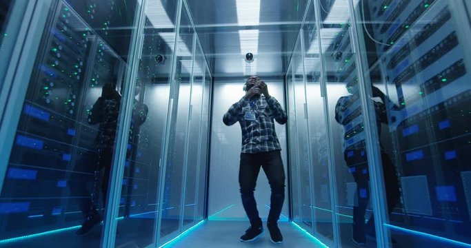 Young African American male IT specialist in headphones dancing in corridor of server room having fun