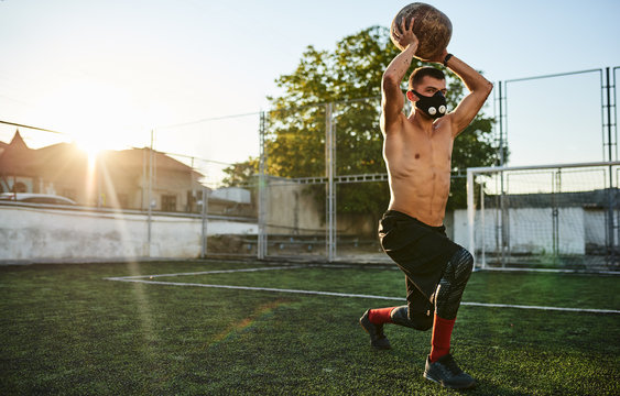Horizontal Shot Of Fit Healthy Muscular Male Doing Exercises With Medicine Ball In Mask In Stadium Outdoors. Copy Space For Advertising Text. Shirtless Sportsman Doing Workout Outside On Sports Ground