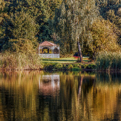 Fototapeta na wymiar Carved wooden white gazebo with red roof by the lake reflected in water in the park, golden hours.