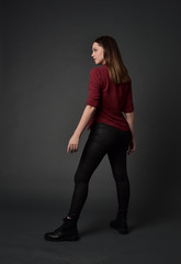 full length portrait of brunette girl wearing  red shirt and leather pants. standing pose, facing away from the camera, on grey studio background.