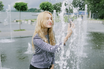 Fototapeta premium Portrait of beautiful student teenager girl holding up a smartphone taking selfies photos, networking using technology, outdoors park. Adolescent with books near water fountain, lifestyle exterior.