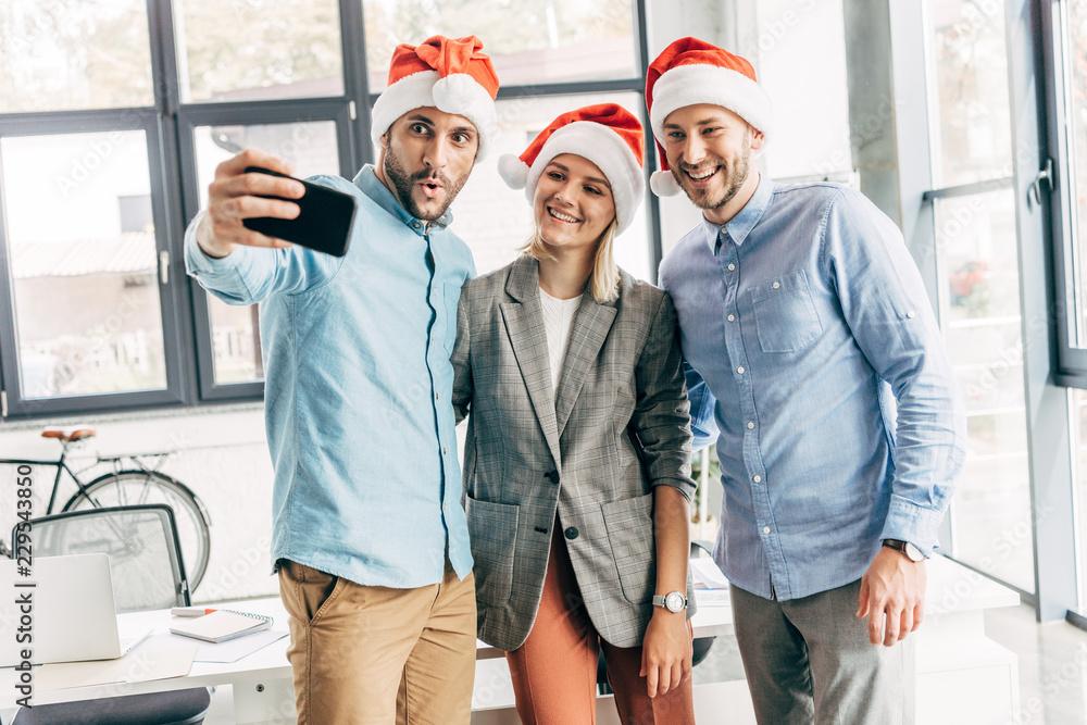 Wall mural happy young start up team in santa hats taking selfie with smartphone in office