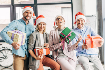 happy coworkers in santa hats holding presents and smiling at camera in office