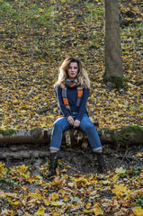 Young beautiful girl with long hair, with a scarf around his neck, sits on a log of fallen tree in the autumn forest among fallen yellow leaves