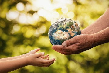 Close up of senior hands giving small planet earth to a child over defocused green background with copy space - obrazy, fototapety, plakaty