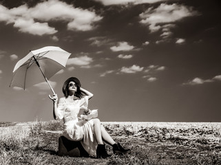 photo of the beautiful young woman sitting on the suitcase with bouquet and umbrella in the field