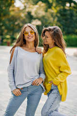 Fototapeta premium Two happy funny hipster girls in bright glasses and pullovers, laughing and sensing kiss, spend free time outside, bright summer outfits and accessories, listening music at headphones.