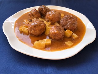 meatballs cooked in sauce of vegetables with a lining of potatoes. Typical Spanish food in white plate and blue background