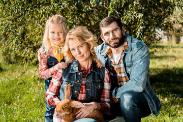 adult farmer family with daughter and brown rabbit sitting on grass outdoors