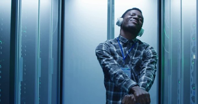 Young African American male IT specialist in headphones dancing in corridor of server room having fun