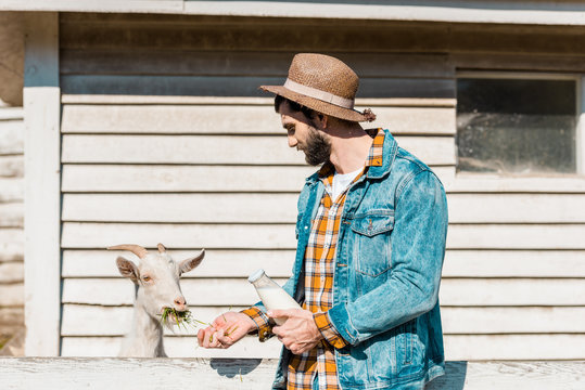 side view of male farmer with bottle of milk feeding goat by grass near wooden fence at farm