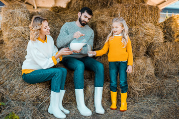adult male farmer pouring milk to little daughter while his wife sitting near at ranch