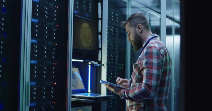 Side view of modern IT engineer using tablet and laptop setting servers in data center while working on mining farm