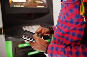 Obraz premium African american man in checkered shirt with backpack holding wallet in hand against atm. Black man traveler.