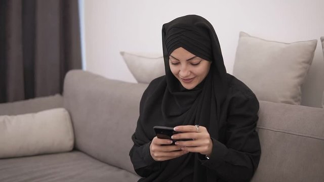 Beautiful muslim woman in black hijab sitting on the sofa and looking to her smartphone and smiling. Modern lofr interior room with white wall on the background