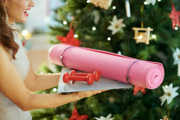 smiling young woman near Christmas tree holding fitness gear