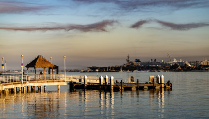 Morning view of sunrise on San Diego Bay from Coronado Island with a pier and dock with an aircraft carrier and cruise ship in the background