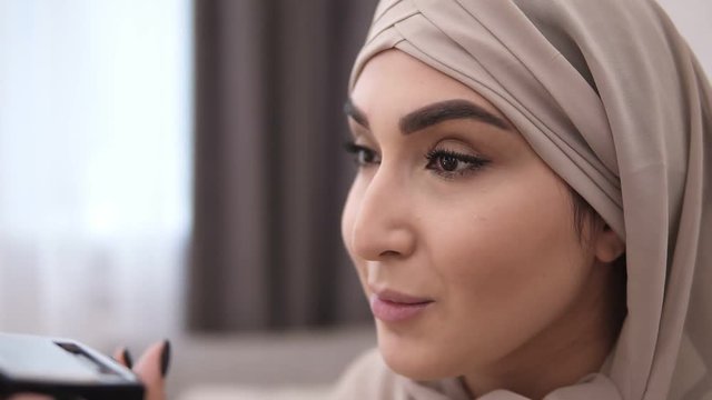Gorgeous muslim woman doing makeup professionally. Brush eyelashes with mascara. Wearing beige headscarf. White wall on the background. Side view