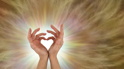 Sending out unconditional love healing vibes - hands making a love heart symbol with a white burst of light behind and a gold brown feather effect background with copy space
