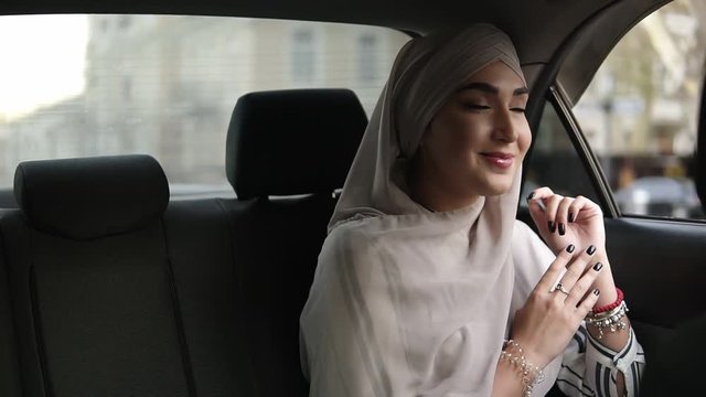 Portrait of a young muslim woman in beige headscarf, sitting in the car while looking out through the window then smiling and posing for the camera. Slow motion