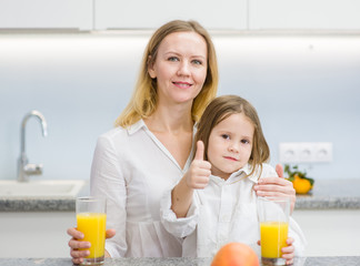 happy family - mother and daughter with orange juice in the kitchen showing thumbs up