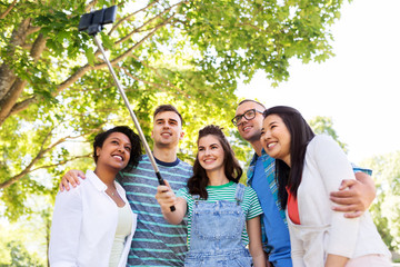 people, technology, friendship and international concept - group of happy friends or students taking picture by smartpnone on selfie stick at summer park