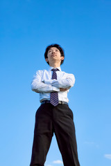 青空をバックに腕組しながら見上げるYシャツ姿の若い男性1人。独立・起業・成功・努力・責任イメージ
