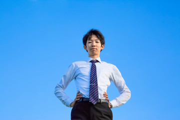 青空をバックに腰に両手を当てカメラ目線の若いYシャツ姿の男性1人立ち姿。ビジネス、成功、野心、向上心イメージ
