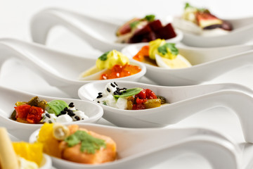 A row of mixed canapes in white ceramic spoons on white background.