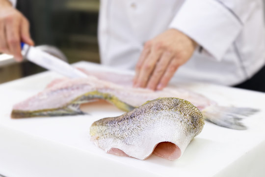 A large zander fish fillet lying on a white chopping board. A chef filleting a fish in the background.