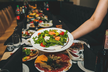 Waitress is holding fresh salad in her hand. Woman sets the table at the restaurant. Cafe service for birthday or wedding celebration. Different dishes on the served table.