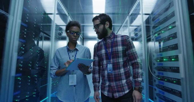 Contemporary multiracial man and woman with tablet walking in server room corridor diagnosing hardware