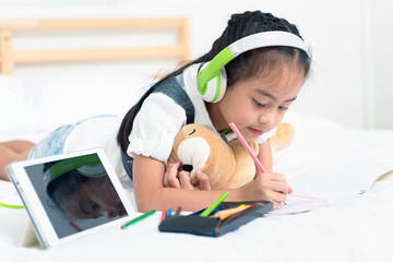 Asian little girl listen to music from tablet with green headphone while writing with colorful pencils on bed in the home. Education for kids and strengthens the imagination of children concept.