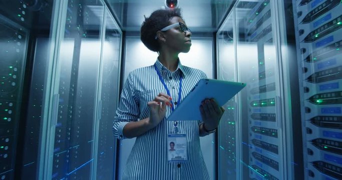 Adult African American woman using tablet while walking among server racks in data center corridor and doing diagnostics