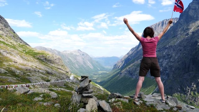 Happy tourist woman arms raised holds norwegian flag, enjoying nature landscape from Trollstigen viewpoint area. Trolls Path mountain road in Norway. Touristic attraction