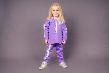 blonde blue-eyed little model girl in a purple sports suit posing in the studio on a black background.active childhood full length portrait violet sporty costume