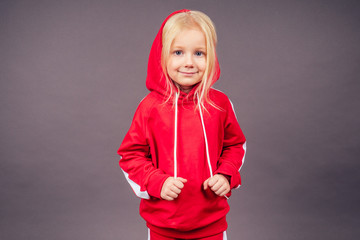 blonde blue-eyed little model girl in a red sports suit posing in the studio on a black background.active childhood full length portrait put on hood on head sporty costume hip-hop dancer