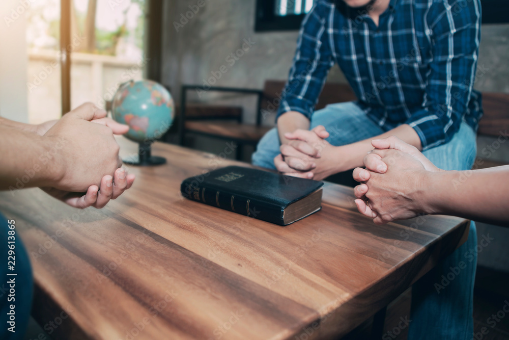 Wall mural Christian friends holding hands together surrounded wooden table with blurred bible and world globe, christian prayer for world mission concept