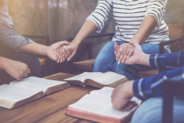 christian small group holding hands and praying together around wooden table with blurred open bible page in home room, devotional or prayer meeting concept - obrazy, fototapety, plakaty