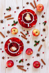 Two cups with hot Christmas fruit drink with spices and berries Christmas background with holly, red apples, cinnamon, anise and cranberries. Top view, close-up on white wooden table