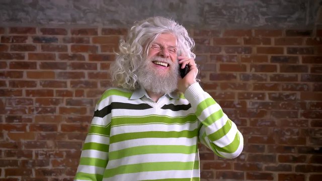 Amusing senior caucasian man with white hair and heavy beard, hanging on his phone and talking with smiley face, happiness illustration, red brick background