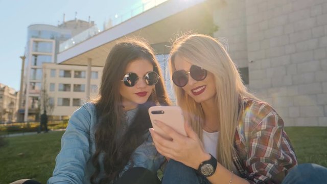 Closeup portrait of two happy hipster girls in casual clothing relax and chat over. Woman using smart phone typing text message outside. They are happy and smiling, sitting on the grass