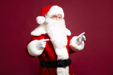 Portrait of man in Santa Clause costume pointing by fingers aside while posing on red background, Christmas and New year concept