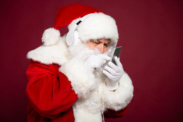 Santa Clause holding smartphone and listening to music at headphone on red background, Christmas and New year concept