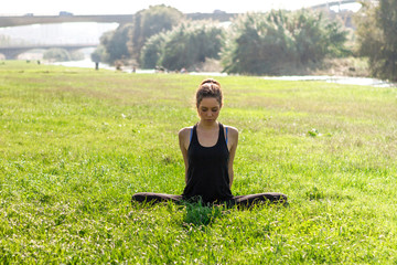 Young athletic woman practicing yoga in a green field with leafy grass next to a river. Young girl practicing postures and yoga exercises with an urban background