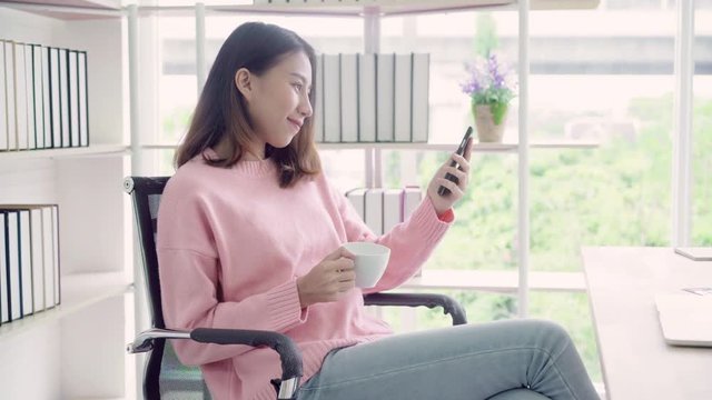 Beautiful smart business Asian woman in smart casual wear using smartphone and drinking warm cup of coffee while sitting on table in creative office. Lifestyle women working at office concept.
