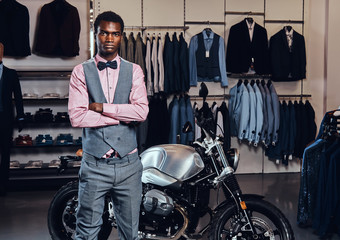 Elegantly dressed African American young man posing with crossed arms near retro sports motorbike at the men's clothing store.