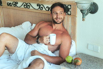 Attractive young bachelor man lying in bed with naked muscular torso and a sexy look with a smirk on his handsome face enjoing his morning. isolated on white background with big biceps and chest