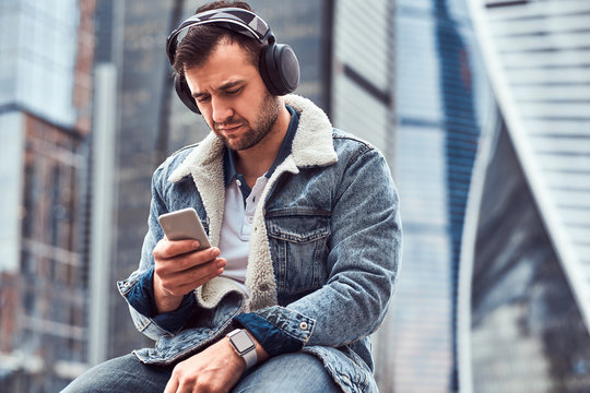 Stylish man wearing denim jacket listening to music and using smartphone sitting in front of skyscrapers in Moskow city at cloudy morning.